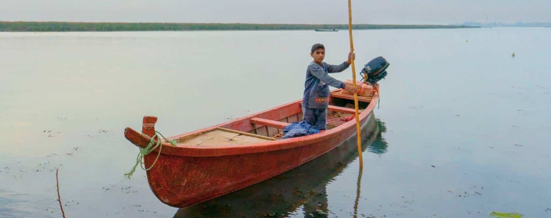 A local Iraqi child rowing a small wooden boat through the Mesopotamian Marshes in southern Iraq, surrounded by reeds and calm waters during a traditional daily life scene.