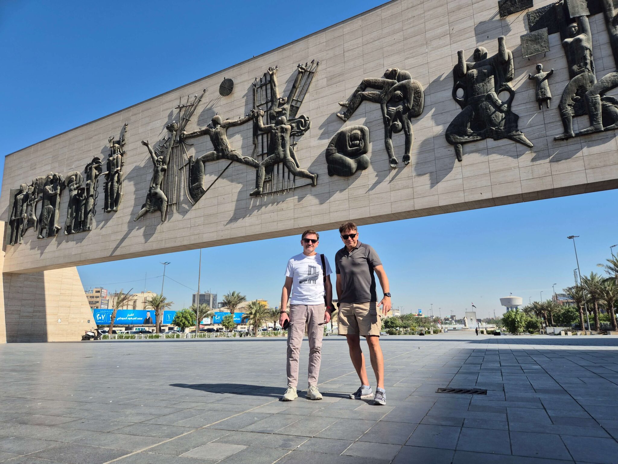 Two tourists standing in Liberation Square, Iraq, with the Freedom Monument in the background during a historical and cultural tour of Baghdad.