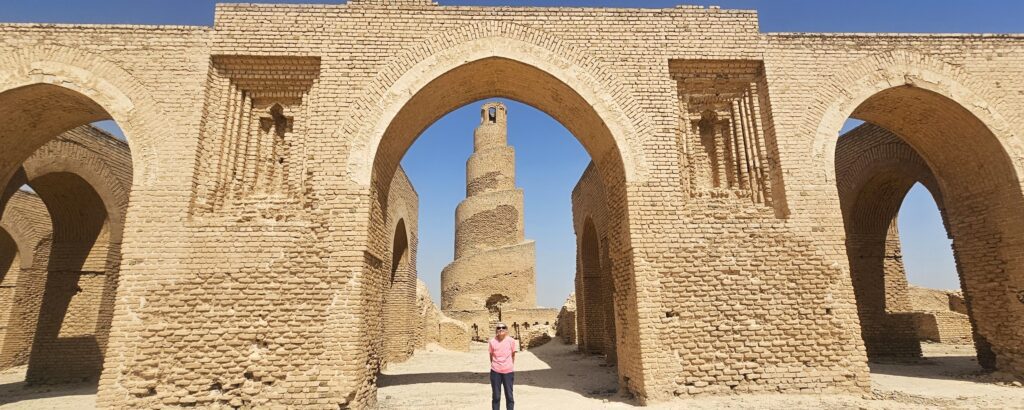 Tourist standing in front of the historic Abu Dulaf Mosque and its distinctive spiral minaret in Samarra, Iraq, with the monumental Abbasid architecture rising prominently across the archaeological landscape.