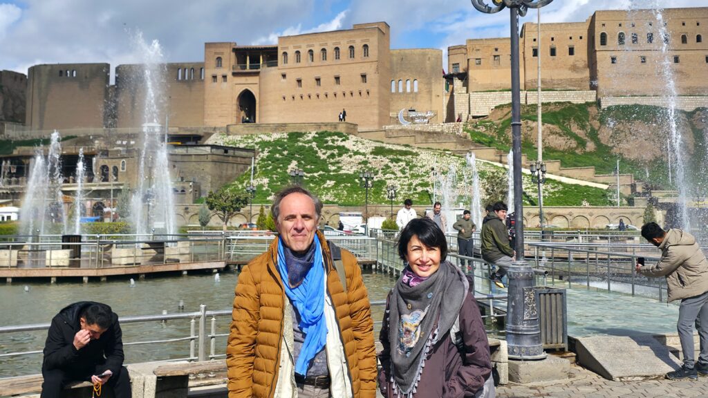 Traveler standing in front of the historic Erbil Citadel in the Kurdistan Region of Iraq, with the ancient elevated settlement, traditional ochre-colored buildings, and distinctive historic skyline visible behind them.